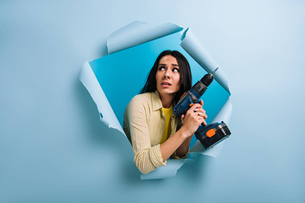 stressed female manual worker with dirty face in paint holding electric drill in torn paper, isolated on blue - Фото, зображення