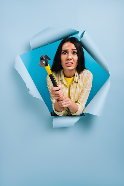 frightened female manual worker with dirty face in paint holding hammer in torn paper, isolated on blue - Photo, image