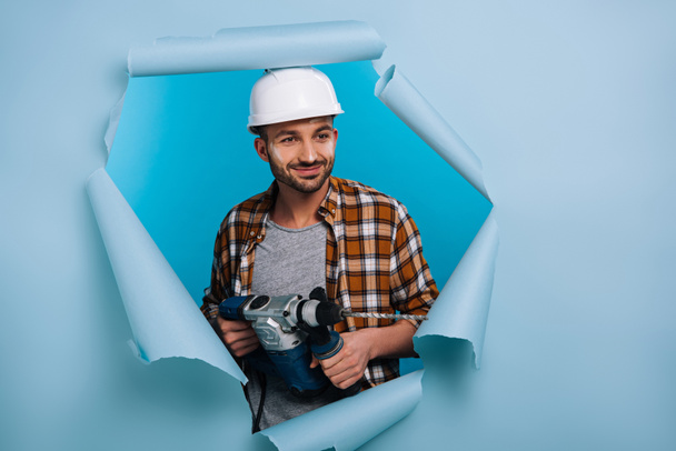 smiling repairman in helmet holding electric drill in torn paper, isolated on blue - Foto, Imagen