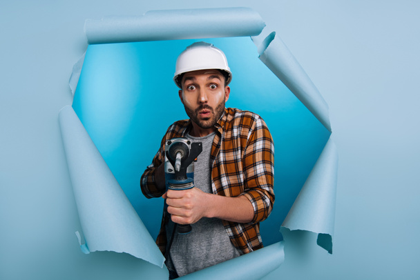 shocked repairman in helmet holding electric drill in torn paper, on blue - Foto, afbeelding
