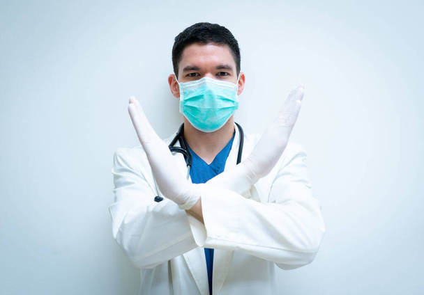 Een jonge knappe dokter in witte jurk en met een masker, met een vingerteken om nee te zeggen tegen geweld in het ziekenhuis. - Foto, afbeelding