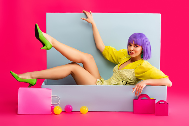 beautiful emotional girl in purple wig as doll lying in blue box with shopping bags and balls, on pink - Valokuva, kuva