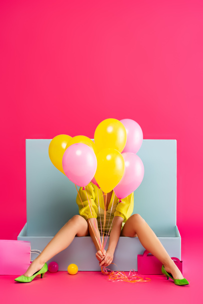 young woman as doll holding balloons and sitting in blue box with balls and shopping bags, on pink - Foto, immagini