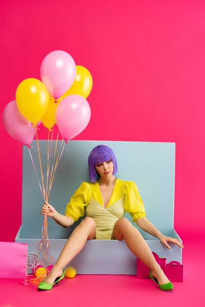 hermosa chica en peluca púrpura como muñeca sosteniendo globos y sentado en caja azul con bolas y bolsas de compras, en rosa
 - Foto, imagen