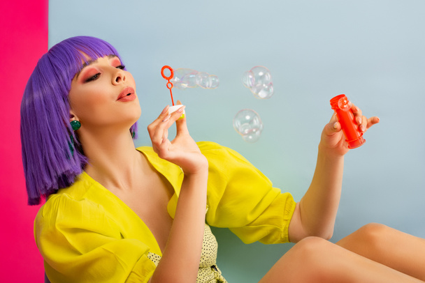 chica atractiva feliz en peluca púrpura como muñeca que sopla burbujas de jabón mientras está sentado en la caja azul, aislado en rosa
 - Foto, Imagen