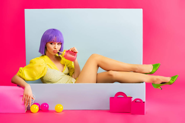 bored girl in purple wig as doll drinking from jar while sitting in blue box with balls and shopping bags, on pink - Photo, Image