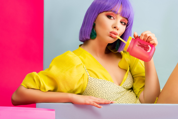 hermosa chica en peluca púrpura como muñeca bebiendo del frasco mientras está sentado en la caja azul, aislado en rosa
 - Foto, Imagen