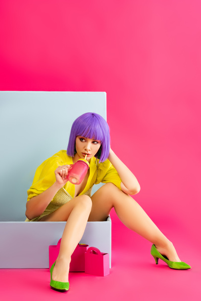 beautiful pop art girl in purple wig as doll drinking from jar while sitting in blue box with shopping bags, on pink - Fotó, kép