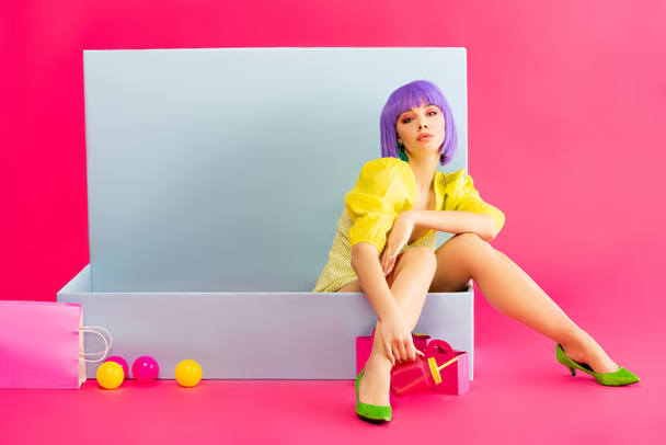 bored girl in purple wig as doll sitting in blue box with balls and shopping bags, on pink - Photo, Image
