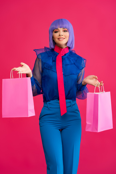 beautiful cheerful girl in purple wig holding shopping bags, isolated on pink - Valokuva, kuva