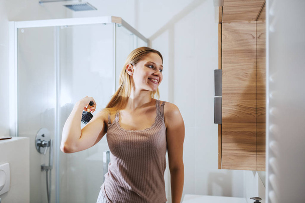 Die junge lächelnde Blondine steht im Badezimmer vor dem Spiegel und kämmt sich die Haare. Morgenroutine. - Foto, Bild