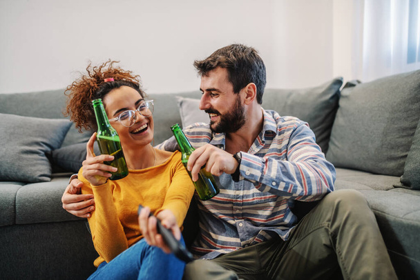 Young attractive couple sitting at home, drinking beer and watching television. Relaxation at sunday afternoon. - Фото, изображение