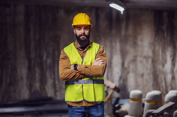 Молодой красивый бородатый рабочий в жилете со шлемом на голове, стоящий внутри тоннеля в процессе строительства с скрещенными руками
. - Фото, изображение