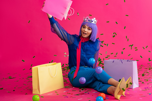 excited girl in purple wig and crown sitting with shopping bags, balls and holiday confetti, on pink - Foto, imagen