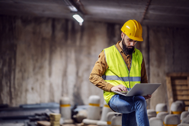 Young attractive focused bearded supervisor in vest with helmet on head standing inside of underground parking lot in construction process and using laptop. - Photo, Image