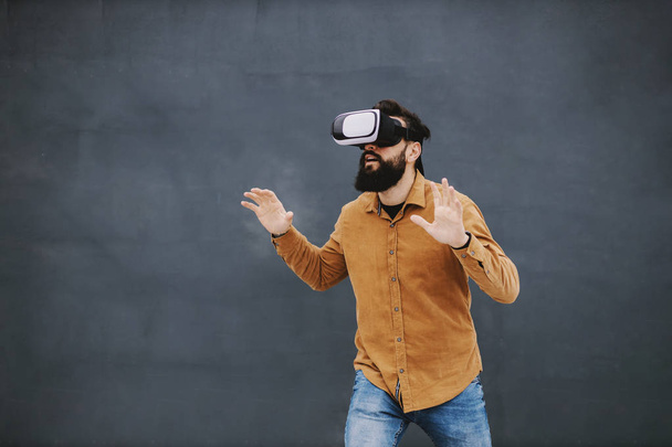 Young attractive bearded hipster standing in front of gray wall and having vr experience. - Φωτογραφία, εικόνα