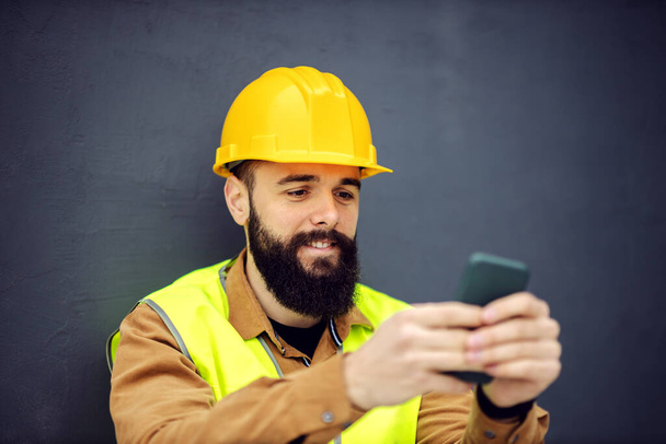 Közelkép jóképű mosolygós szakállas munkás mellényben, sisakkal a fején ül, okos telefont használ, és szünetet tart a munkában. - Fotó, kép