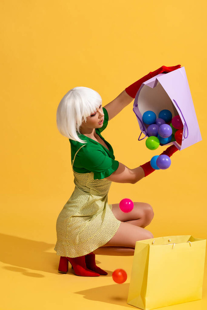 surprised girl in white wig holding shopping bags with colorful balls, on yellow - Photo, Image