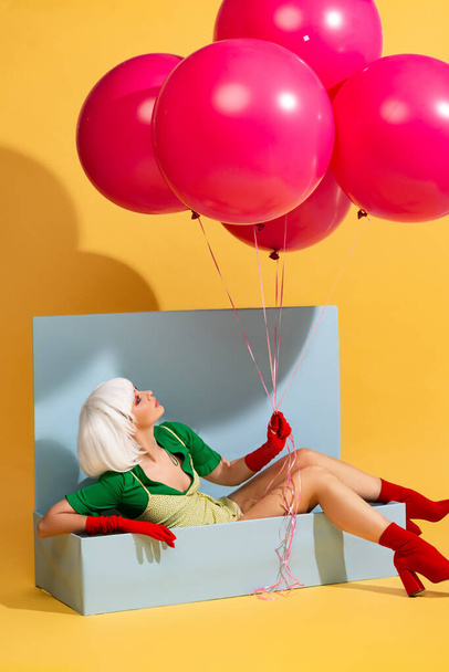 attractive pop art girl in white wig as doll sitting in blue box and holding balloons, on yellow - Foto, Imagem
