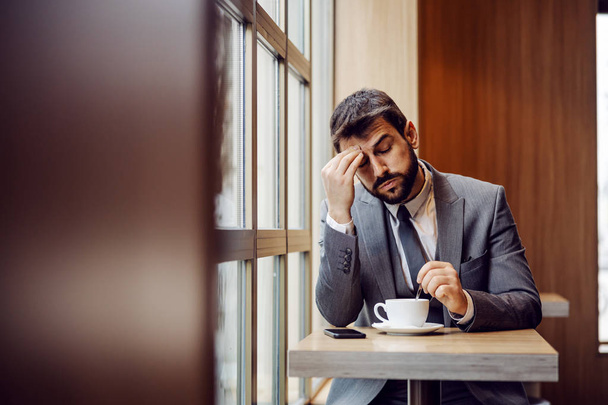 Der junge bärtige Geschäftsmann sitzt im Café neben dem Fenster und hat Kopfschmerzen. Er glaubt, Kaffee am Morgen könnte helfen. - Foto, Bild