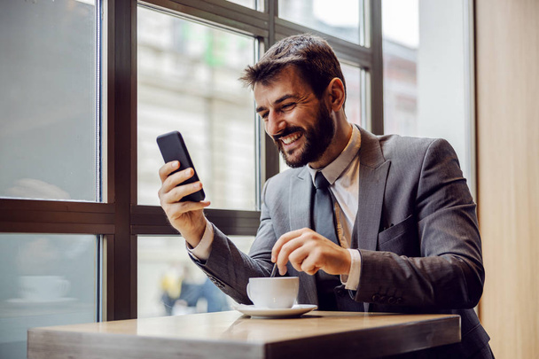 Giovane uomo d'affari barbuto sorridente seduto in caffetteria, leggendo qualcosa di divertente su smart phone e mescolando il caffè
. - Foto, immagini