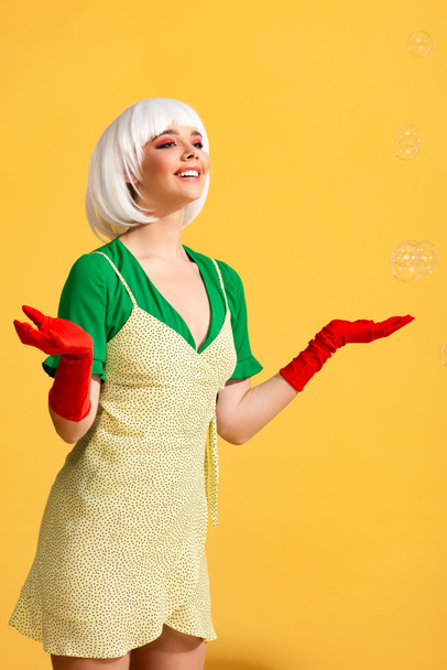 attractive smiling pop art girl in white wig with soap bubbles, on yellow  - Valokuva, kuva