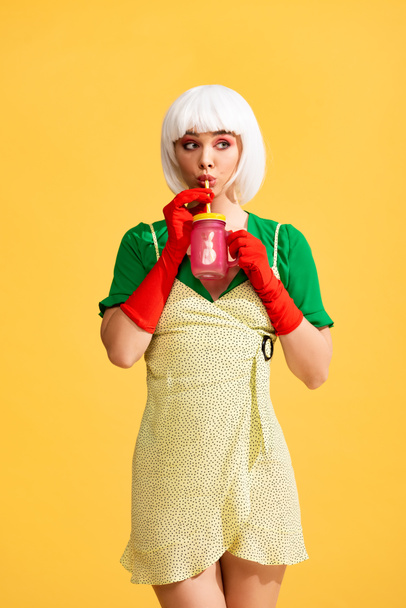 surprised pop art girl in yellow dress and white wig drinking fresh beverage from jar, isolated on yellow - Photo, Image