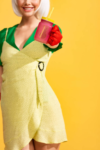 cropped view of smiling pop art girl in yellow dress holding jar with fresh drink, isolated on yellow - Photo, Image