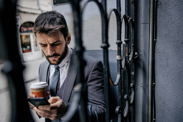 Young bearded businessman standing next to an old iron fence in font of building, holding cup with fresh morning coffee and typing message on smart phone. - Fotografie, Obrázek
