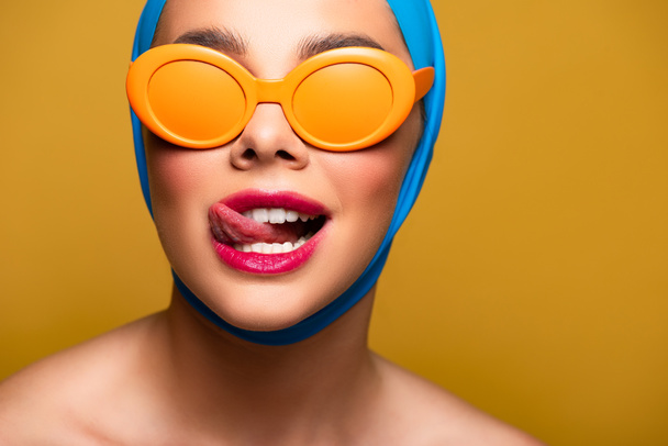 happy girl in scarf and fashionable sunglasses licking lip, isolated on yellow - Valokuva, kuva