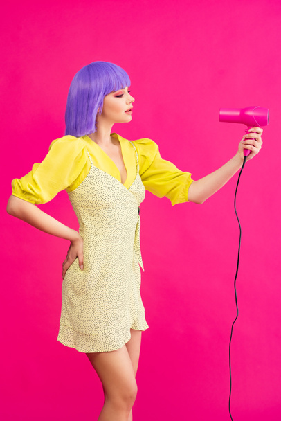 fashionable pop art girl in purple wig using hair dryer, isolated on pink - Foto, imagen