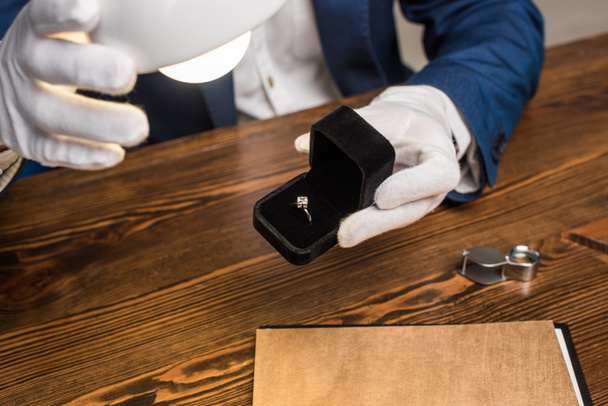 Vista recortada del anillo de sujeción del tasador de joyas con gemas en caja cerca de la lámpara en la mesa aislada en gris
 - Foto, Imagen
