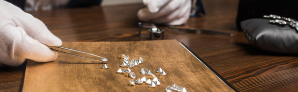 Vista recortada del tasador de joyas con pinzas que examinan las piedras preciosas en la mesa de madera aislada en gris, tiro panorámico
 - Foto, imagen