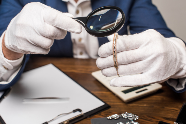 Ausgeschnittene Ansicht des Schmuckgutachters untersucht Halskette mit Lupe in der Nähe von Taschenrechner, Klemmbrett und Ergebnis auf Tisch isoliert auf grau - Foto, Bild