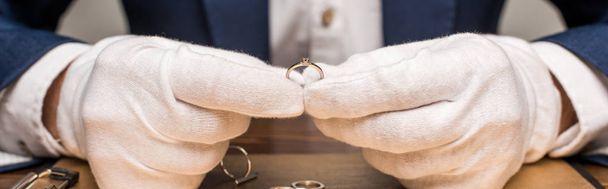 Vista recortada del tasador de joyas que sostiene el anillo de joyería cerca de bordo en la mesa aislada en gris, tiro panorámico
 - Foto, Imagen