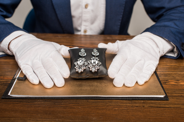 Cropped view of jewelry appraiser holding jewelry earnings on jewelry pillow near board on table isolated on grey - Fotografie, Obrázek