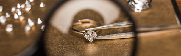 Selective focus of magnifying glass, jewelry ring with gemstone in tweezers on board, panoramic shot - Photo, Image
