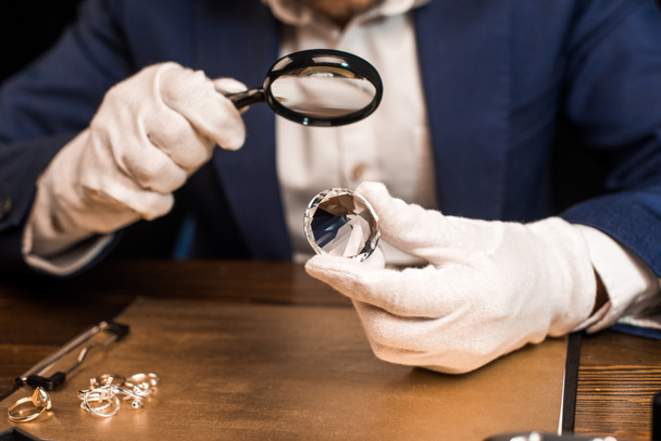 Vista recortada del tasador de joyas que sostiene piedras preciosas y lupa cerca de anillos de joyería a bordo en la mesa aislada en negro
 - Foto, imagen