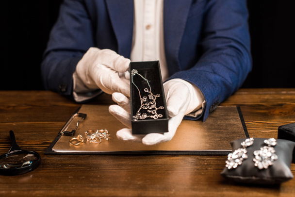 ブラックで隔離されたテーブルの上にボード上の宝石類の近くのネックレスと宝石鑑定人保持ボックスの作物ビュー  - 写真・画像