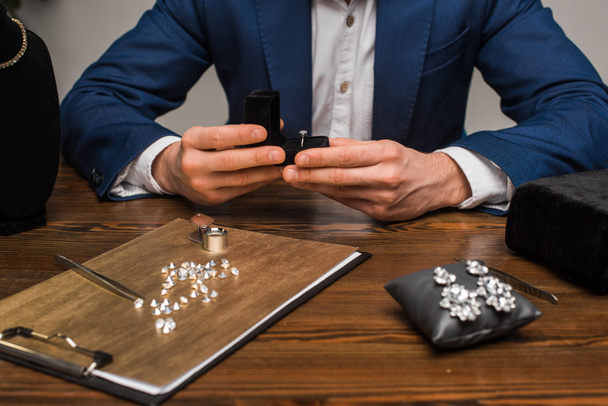 Περικοπή άποψη του εκτιμητή κοσμήματα κρατώντας κουτί με δαχτυλίδι κοσμήματα κοντά κοσμήματα και εργαλεία στο τραπέζι σε γκρι φόντο - Φωτογραφία, εικόνα