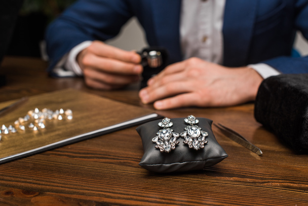 Selective focus of earrings on jewelry pillow and jewelry appraiser working at table in workshop - 写真・画像