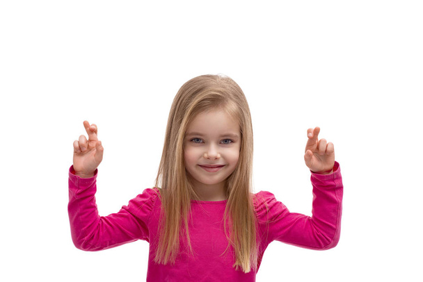 Περιμένετε μέχρι το πορτρέτο ενός χαριτωμένο ξανθιά κοπέλα με μακριά μαλλιά με τα δάχτυλα σταυρωμένα κάνοντας μια ευχή σε λευκό φόντο στο στούντιο. Ευτυχισμένη παιδική ηλικία  - Φωτογραφία, εικόνα