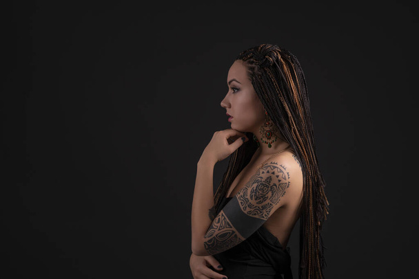 Half length portrait of a young woman with a  polynesian  tattoo on her arms and with african pigtails against  dark background with space in the studio - Photo, Image
