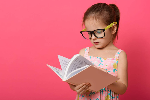 Image of cute preschooler girl wearing sundress and eyewear, female kid reading textbook while standing isolated over rose studio background, child enjoying to read interesting fairy tale. Education. - Zdjęcie, obraz