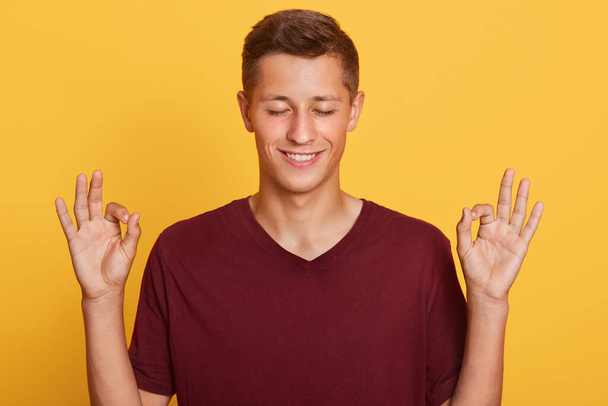 Horizontal indoor picture of peaceful positive handsome guy closing his eyes, smiling sincerely, making gesture, showing sign okay, wearing dark red t shirt, standing isolated over yellow background. - Photo, image