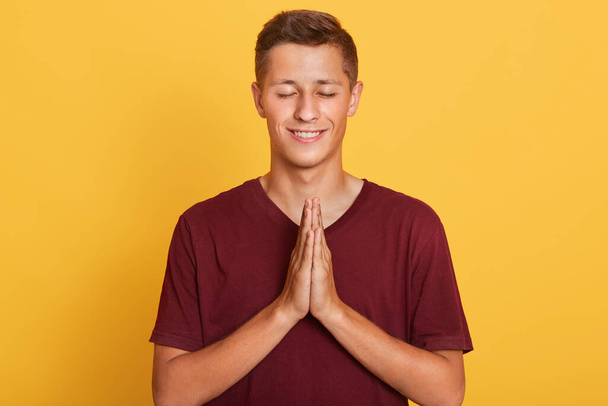 Image of energetic good looking fair haired young boy closing his eyes, putting hands together, making peaceful gesture, relaxing, meditation time, wearing red t shirt. People and emotions concept. - Foto, Bild