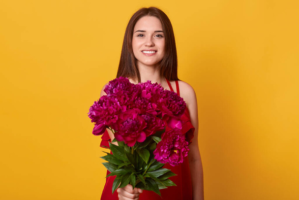Horizontal shot of cute young woman with flowers standing isolated over yellow background, female giving bouquet of maroon peonies to somebody, brunette girl with flowers in hands. Holiday concept. - Foto, imagen