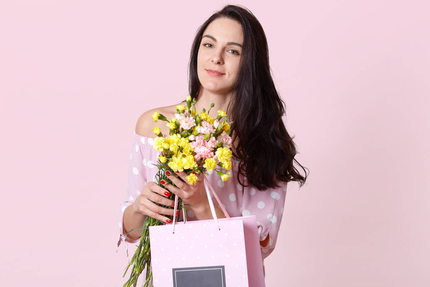 Image of charismatic beautiful attractive young brunette looking directly at camera, standing isolated over light pink background, holding flowers and present in package. Womens day concept. - Foto, Bild