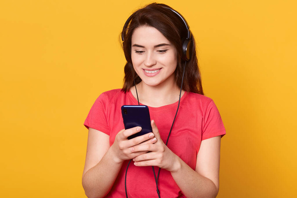 Indoor shot of young girl listening and enjoying music, holding cellular phone in hands and earphones on top of her head, girl wearing red casual t shirt, model posing isolated over yellow background. - Photo, image