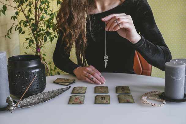 beautiful young woman in a black velvet dress holds a crystal on a silver chain above the runes during a spiritual seance - Фото, изображение